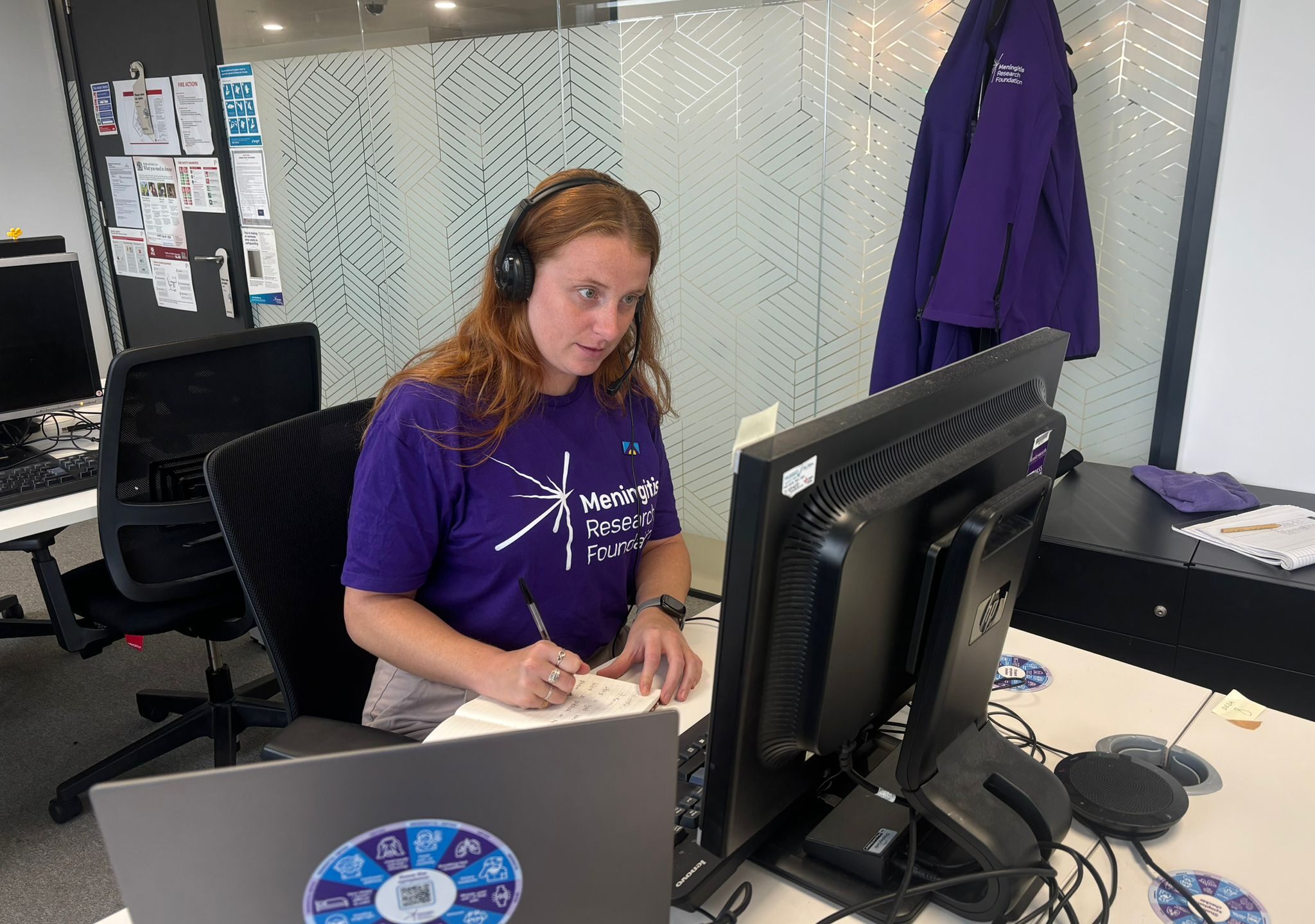 Person speaks on the phone at a desk wearing a Meningitis Research Foundation t-shirt.