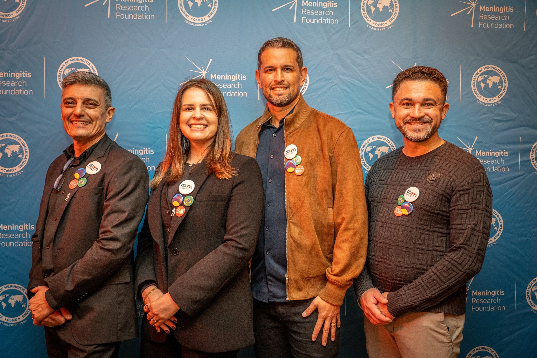 Three men and one woman standing in a row, dressed smartly and smiling at the camera. They are standing in front of a blue backdrop with the Meningitis Research Foundation and CoMO logos and they are all wearing a number of pin badges, including one which says 