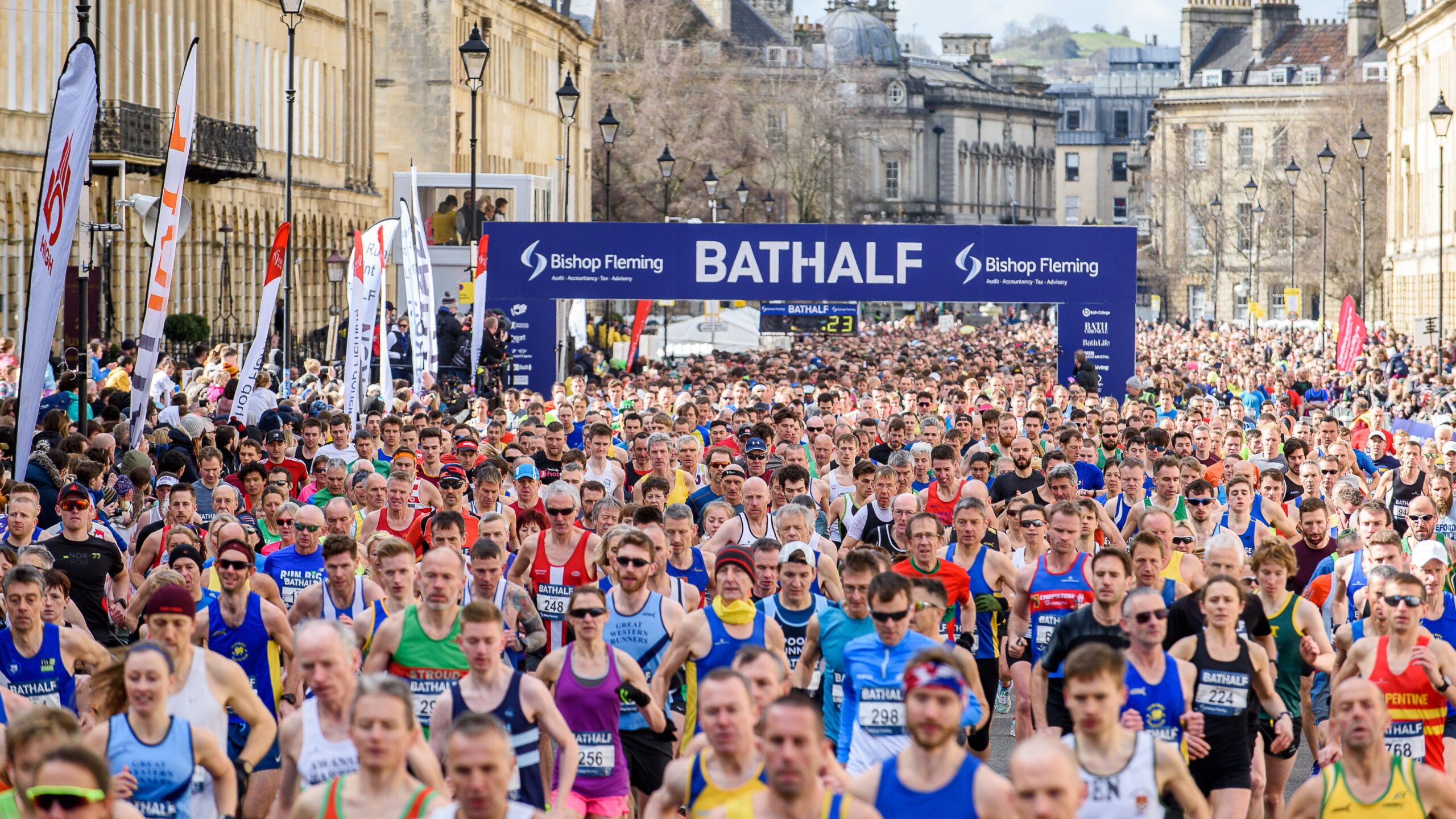 Crowd of people running down a street in front of a Bath Half Marathon start sign.