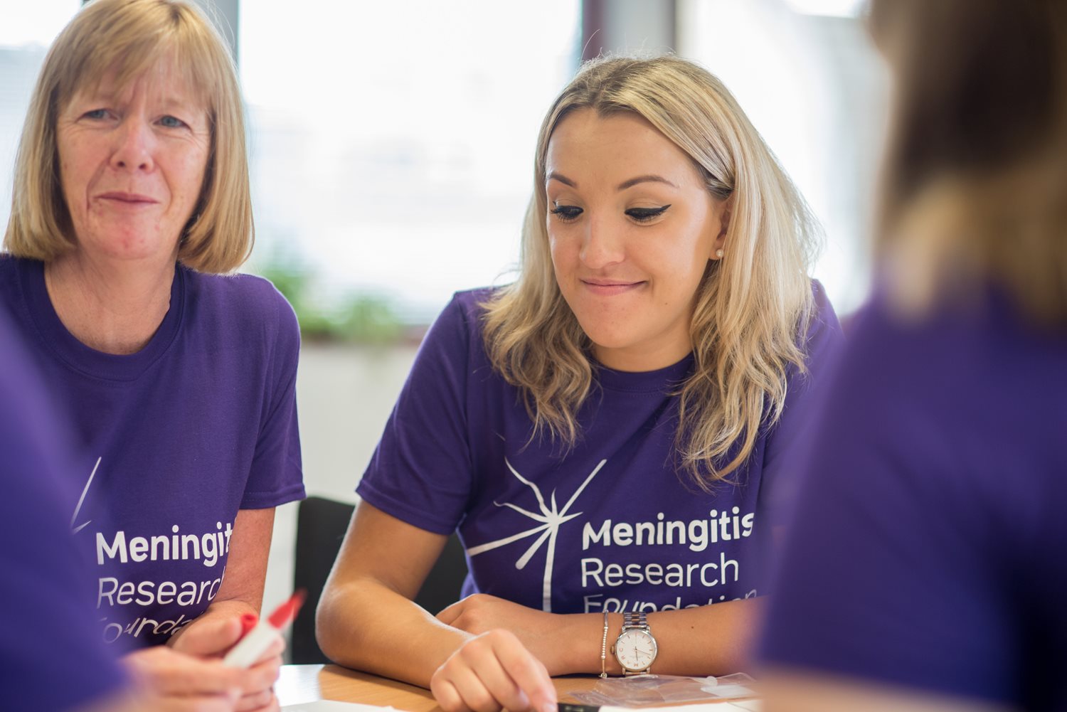 Two women sitting at a table wearing Meningitis Research Foundation t-shirts smiling.