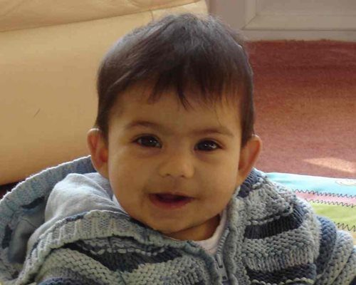 A photograph of a baby girl with brown hair wearing a cardigan smiling.