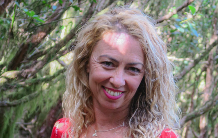A woman with blonde hair and a red top, standing in front of a tree and smiling.