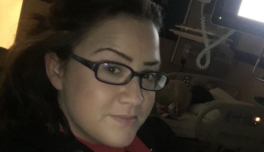 A woman with brown hair and glasses in front of a hospital bed with a sleeping child.