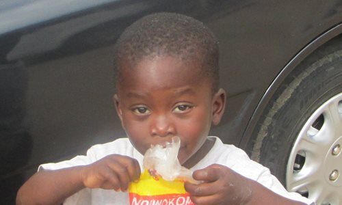 A young boy standing in front of a car eating from a bag of snacks.