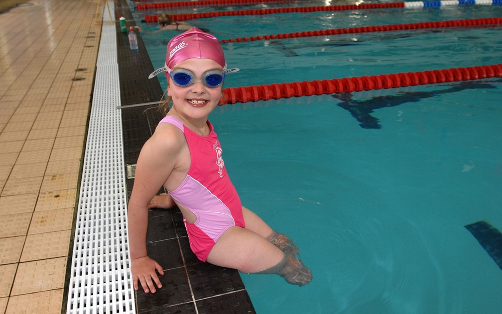 A young girl with lower limb loss dangling her legs in a swimming pool and smiling.