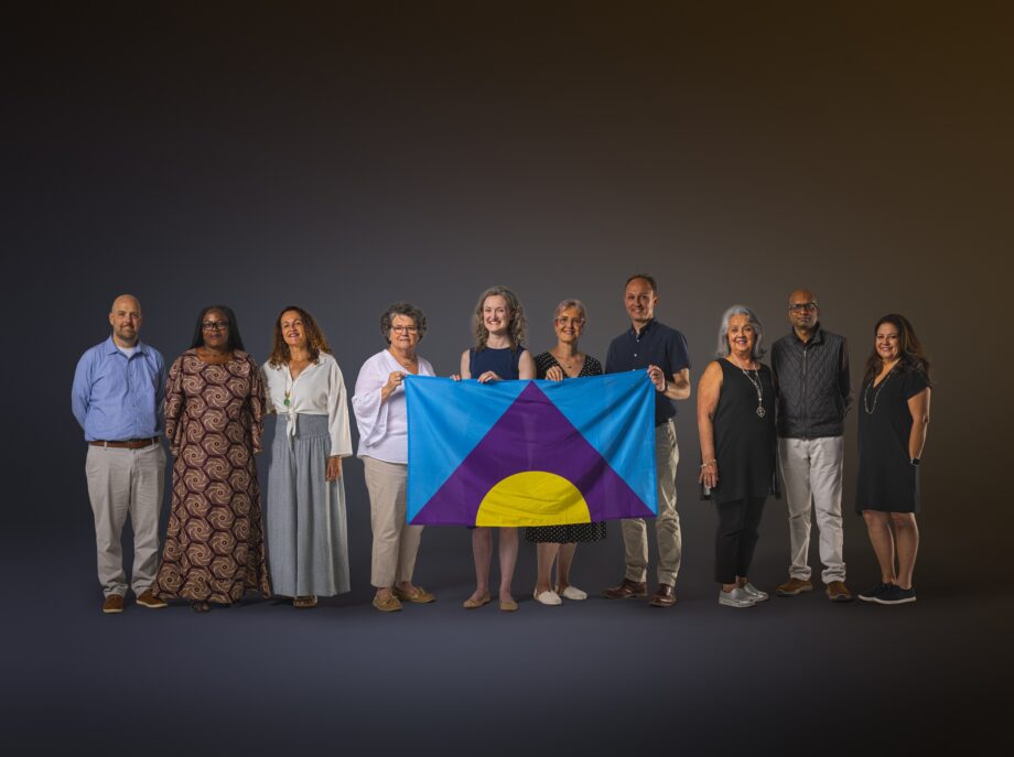 Group of people stood in a line in front of a grey background, holding the Meningitis Flag whilst facing the camera and smiling.