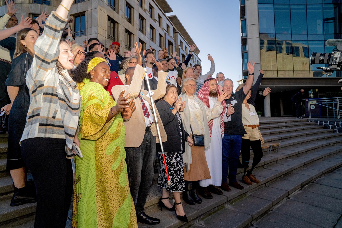 Big group of people from many different countries stood outside two buildings, on steps. All have their hands in the air and are smiling, waving and cheering at the camera.