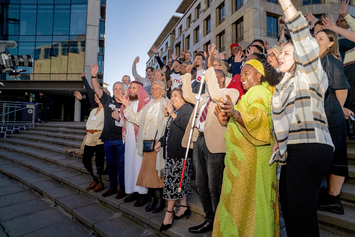 Big group of people from many different countries stood outside two buildings, on steps. All have their hands in the air and are smiling, waving and cheering at the camera.