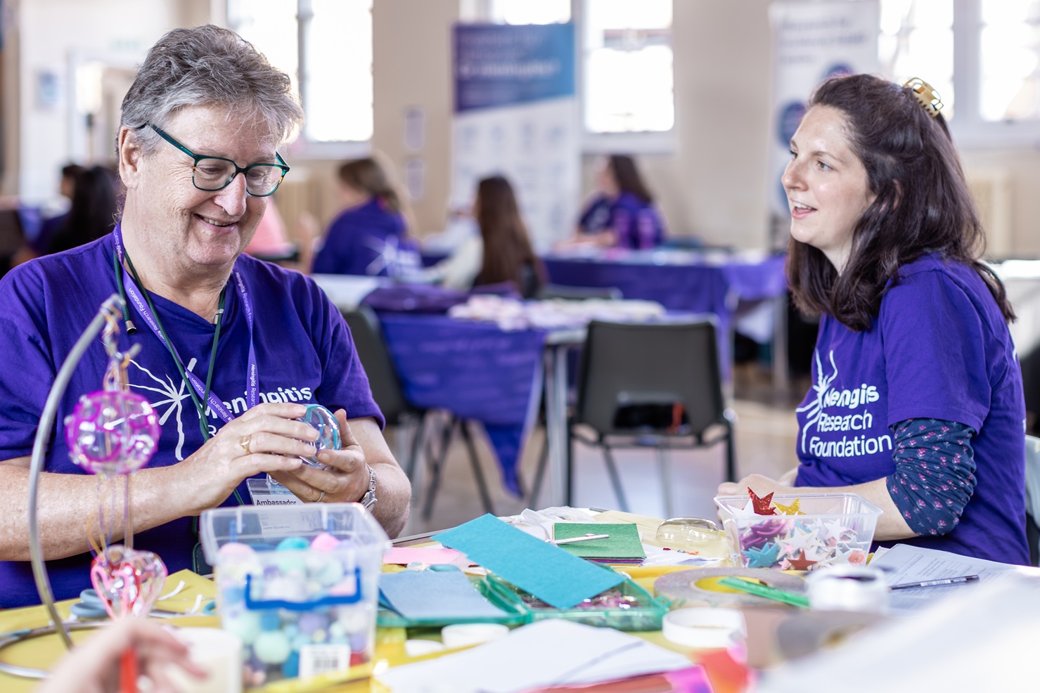A man and a woman sat at a table together whilst the man is crafting. The woman is talking and the man is smiling.