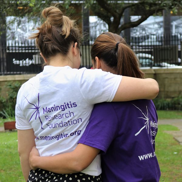 Two women wearing Meningitis Research Foundation t-shirts, one in purple and one in white, have their arms around eachother, with their backs facing the camera. They are in a park and there is grass and trees in the background.