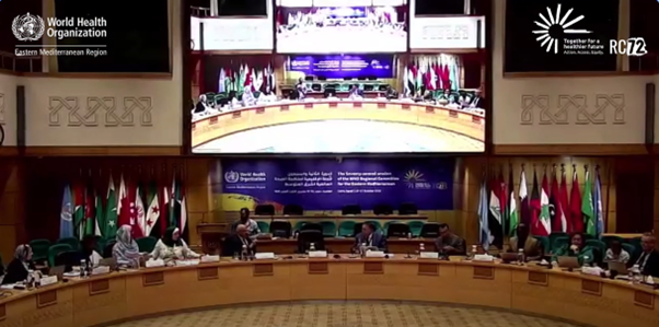 An official meeting with a number of people sitting at a crescent-shaped table with microphones in front of them, flags hanging in the background and a big screen behind them.