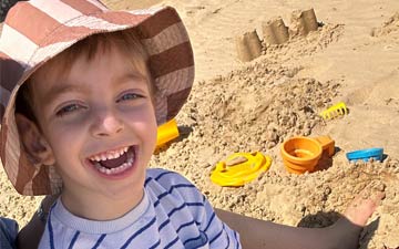 A young boy sitting on the beach in front of some sandcastles, smiling.
