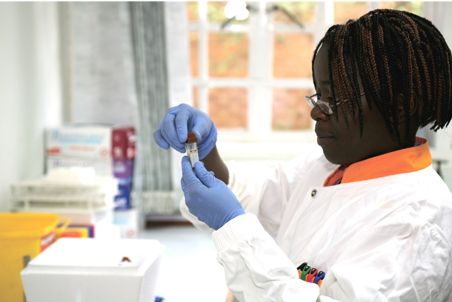 Woman in a lab, holding and looking at a test tube, wearing a lab coat and gloves.