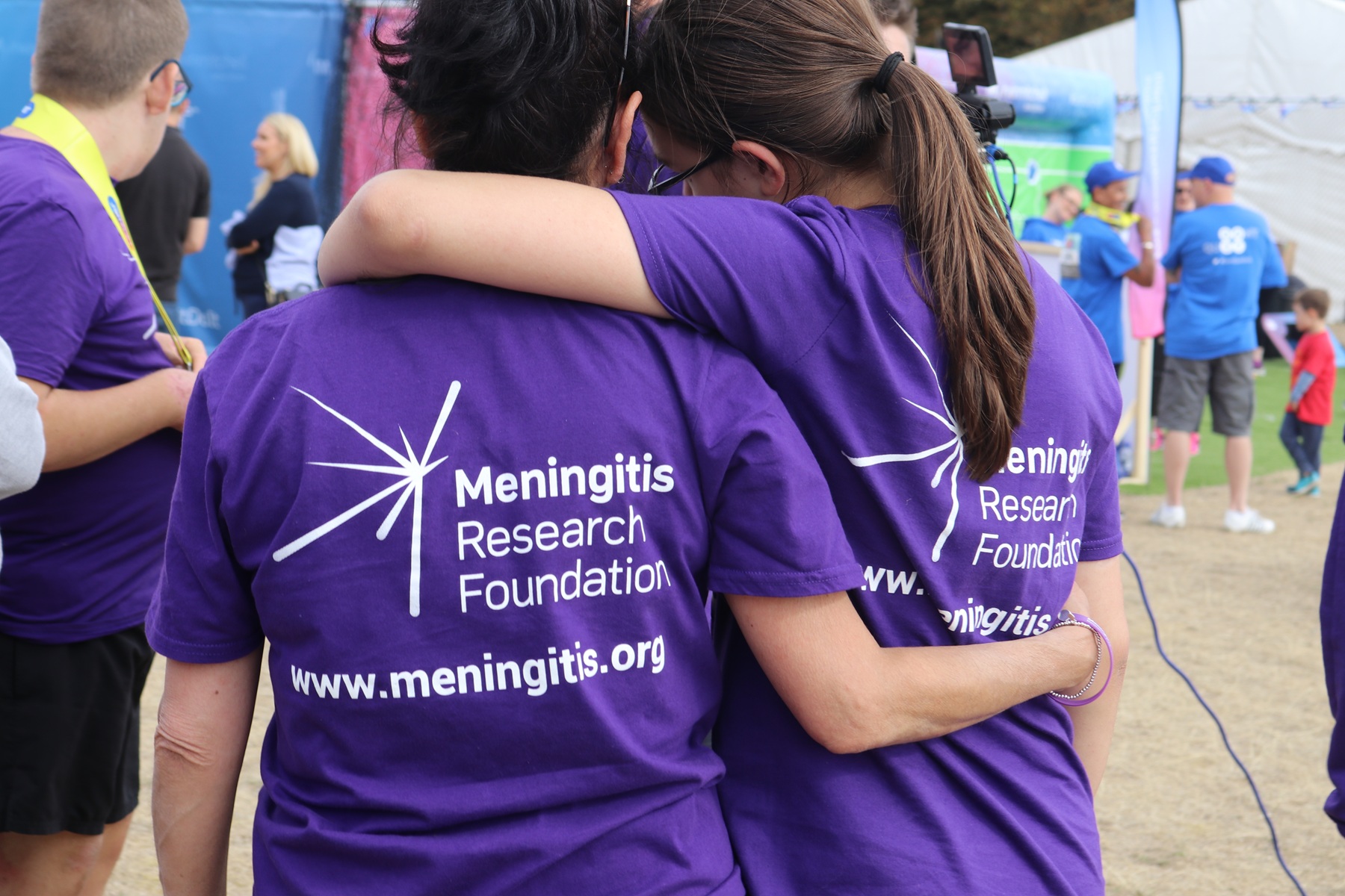 Two women wearing Meningitis Research Foundation t-shirts embracing, with their backs to the camera.