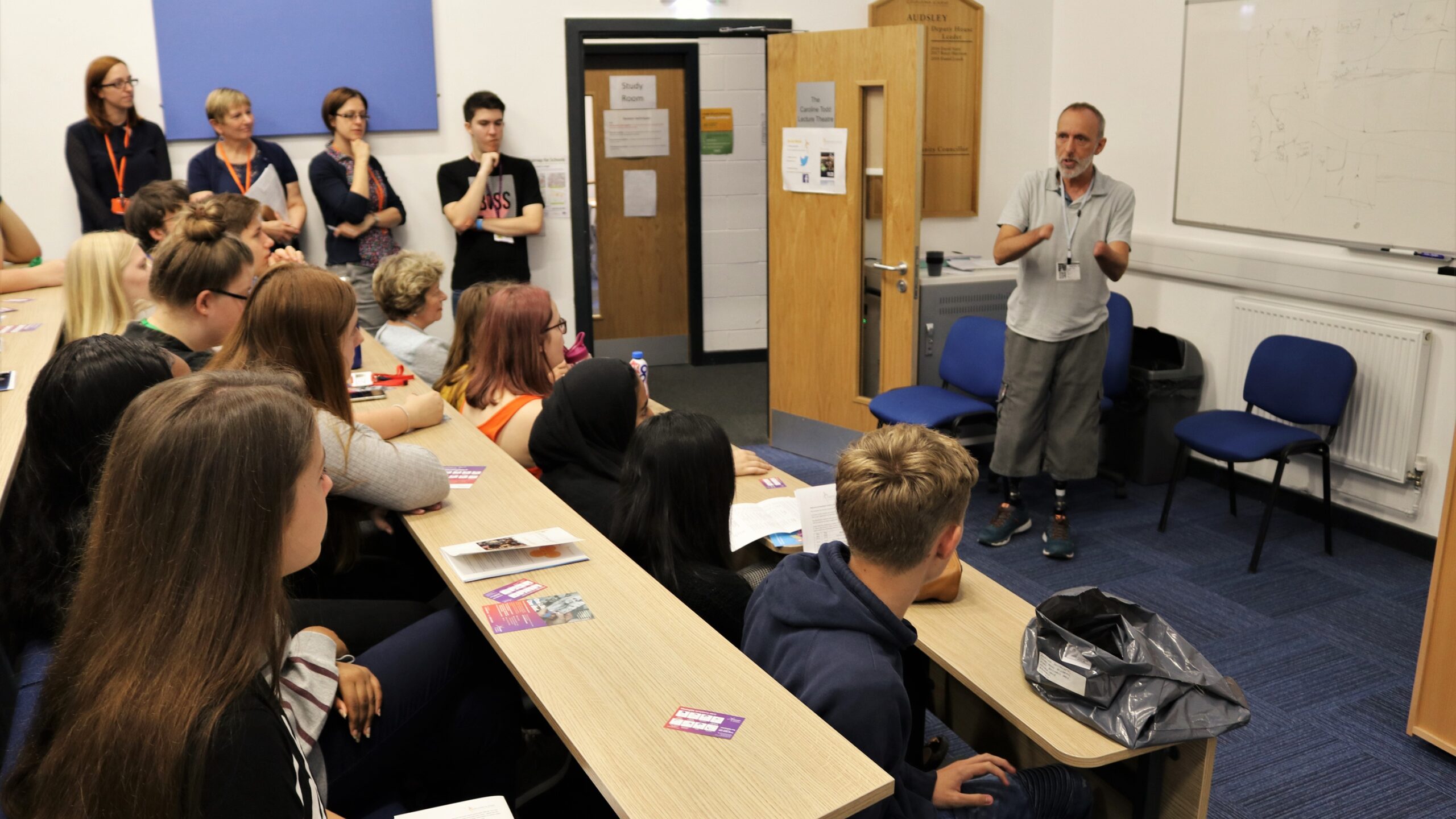 Classroom of teenagers sat at tiered desks listening to a man who has survived meningitis talking about his experience. Man is at the front of the teenagers standing up and showing his prosthetic legs.