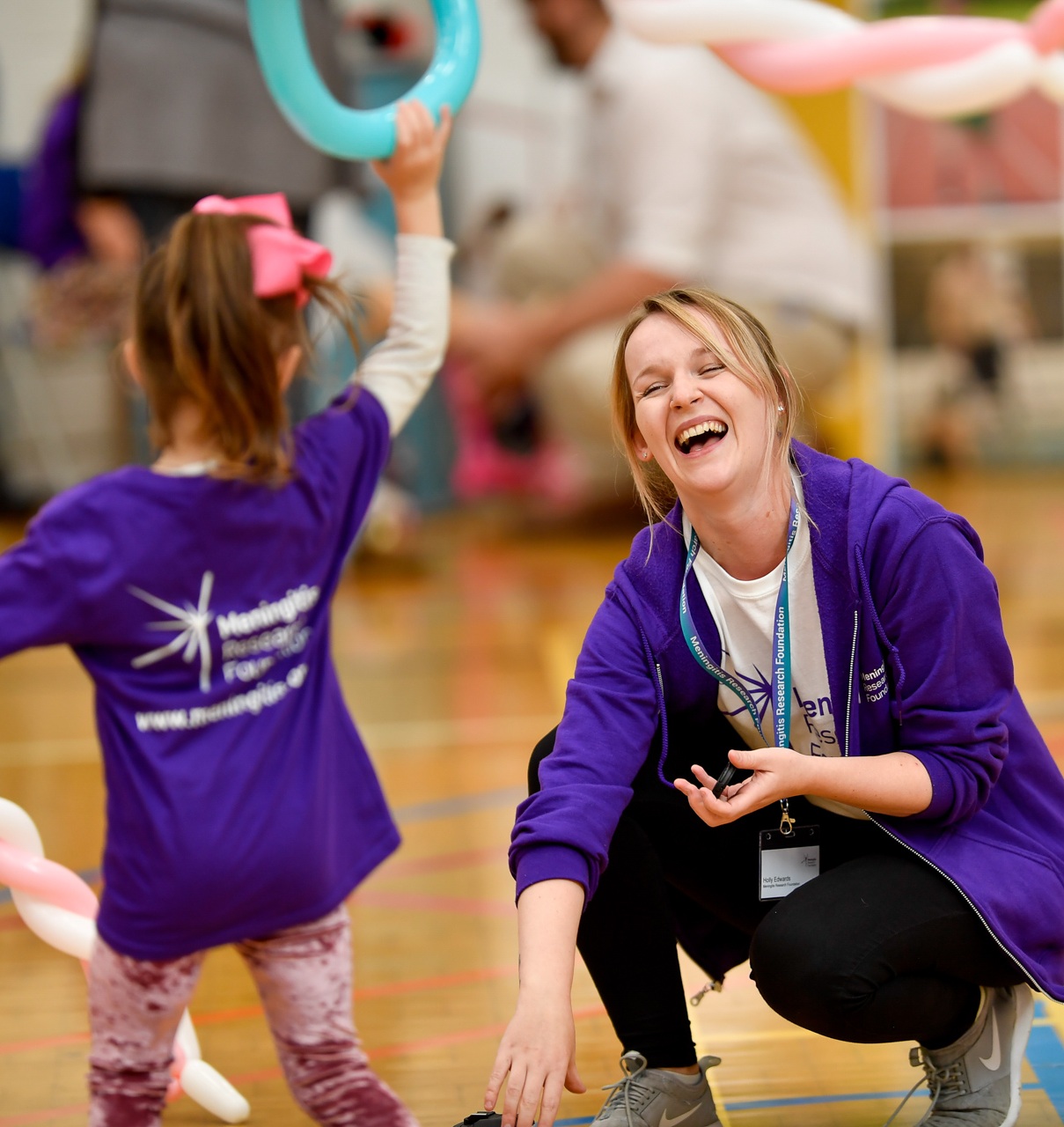 Child wearing a Meningitis Research Foundation t-shirt holds a blue balloon in the air. An adult in a Meningitis Research Foundation t-shirt is crouched down and smiling with them