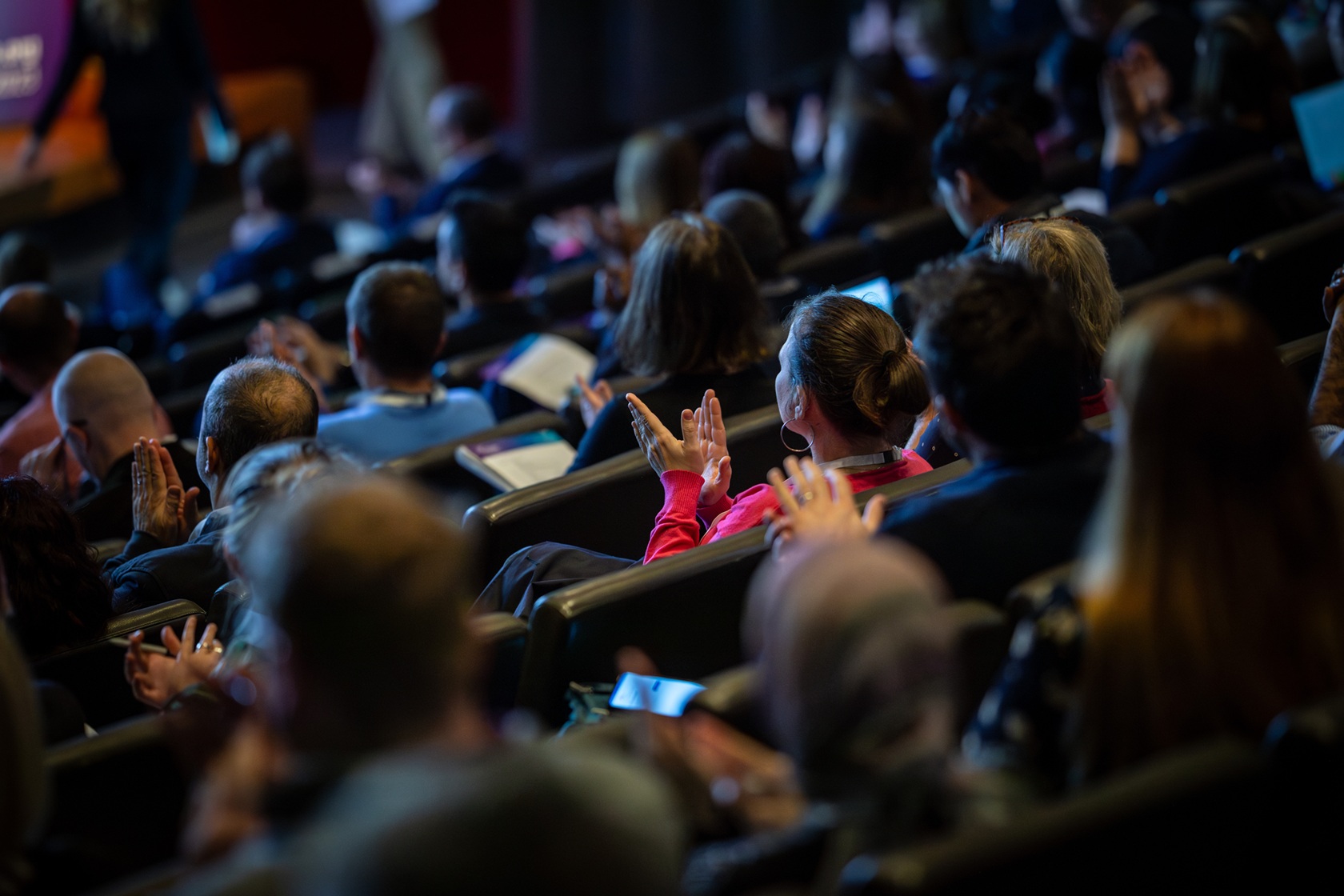 Rows of people seated and clapping at an event, taken from behind so you are seeing the back of them.
