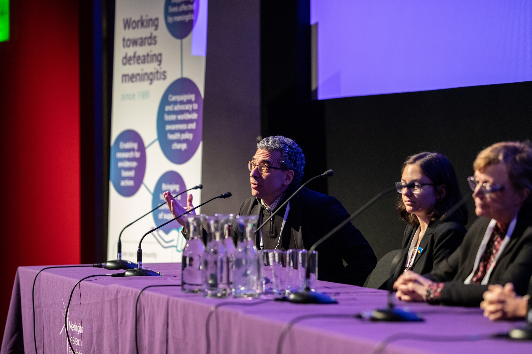 Man at an event, sat at a table speaking into a microphone, with two women sat next to him.