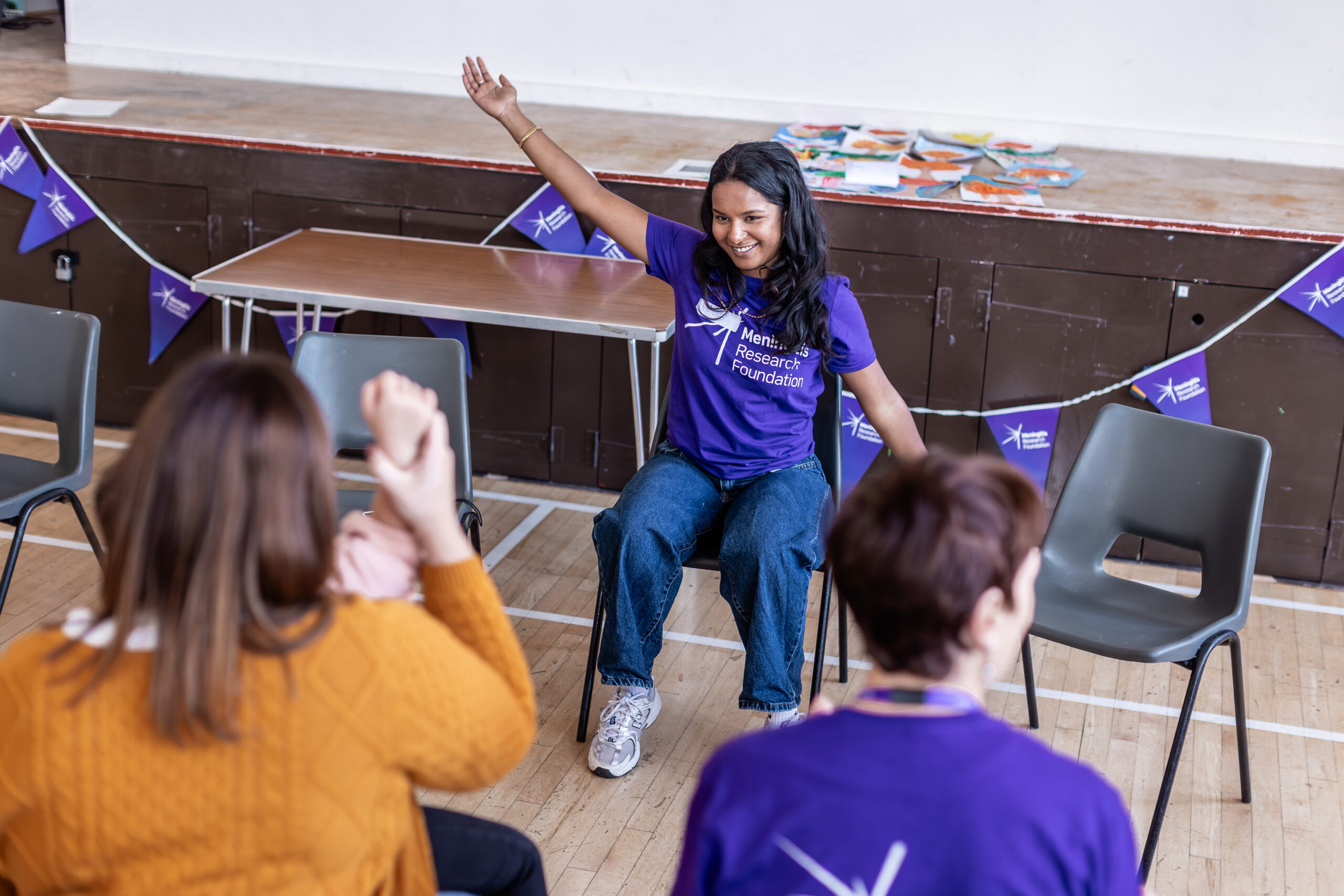 A woman in a purple t-shirt is demonstrating seated exercises to some people sat in front of her