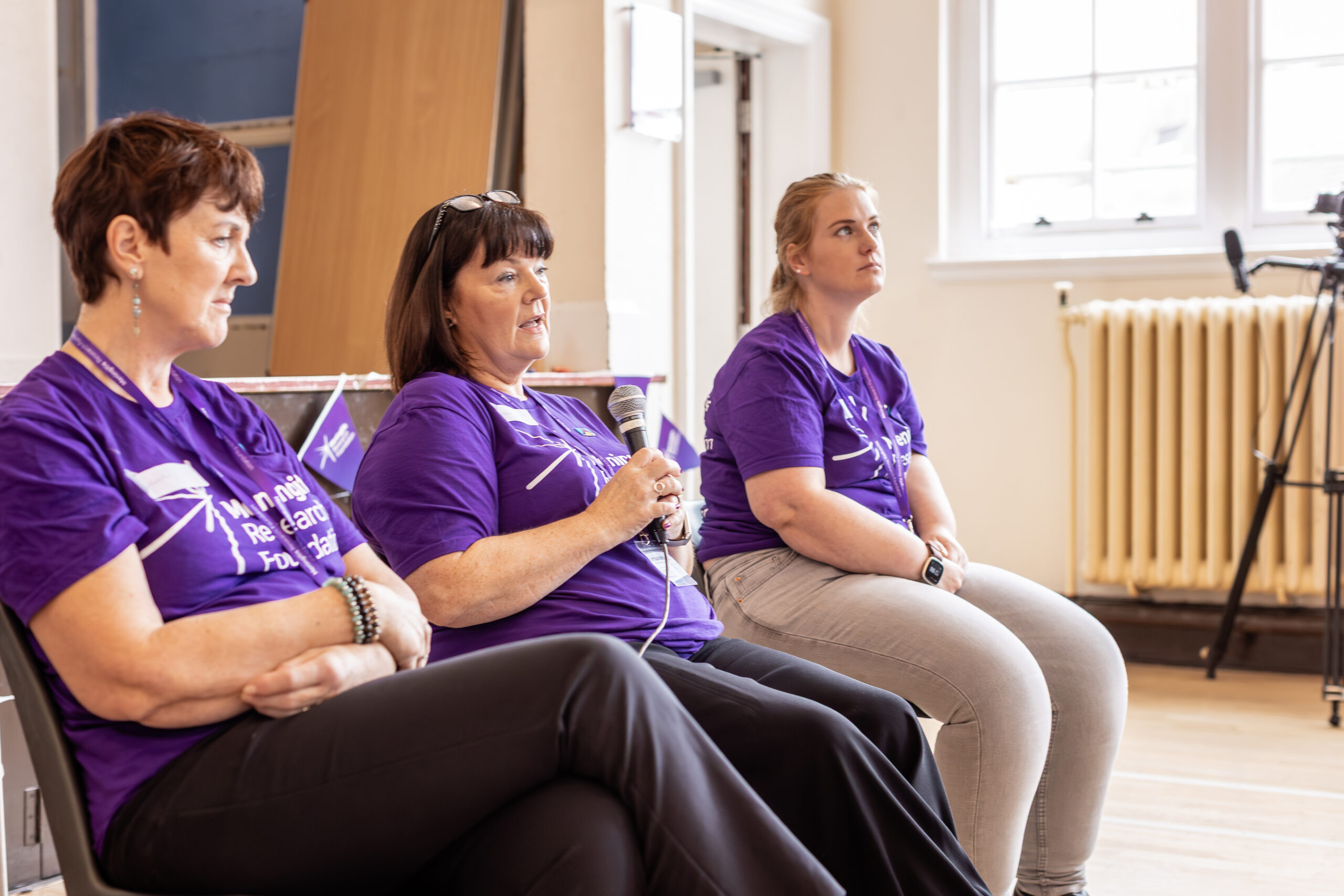 Three women sat in a line, with the middle one speaking into a microphone