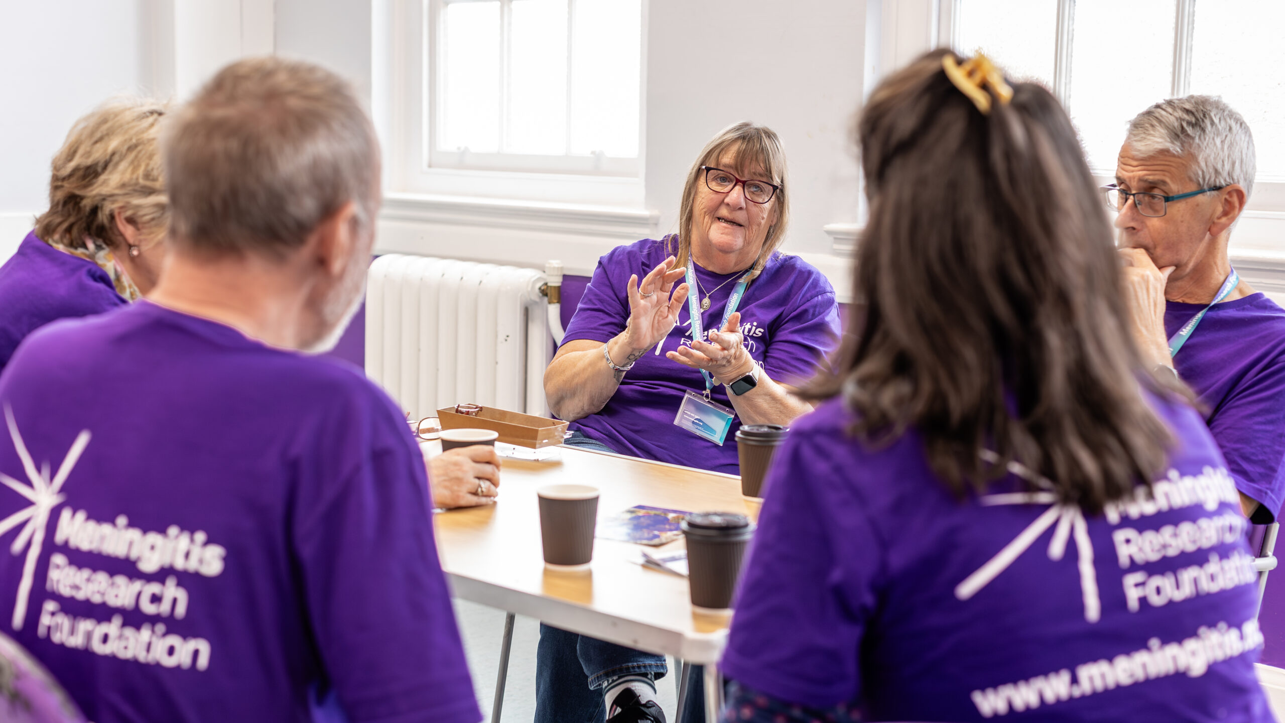 Woman speaking to a group around her, all in purple Meningitis Research Foundation t-shirts