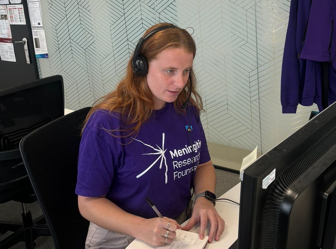 A person speaking on the phone at a desk wearing a Meningitis Research Foundation t-shirt.