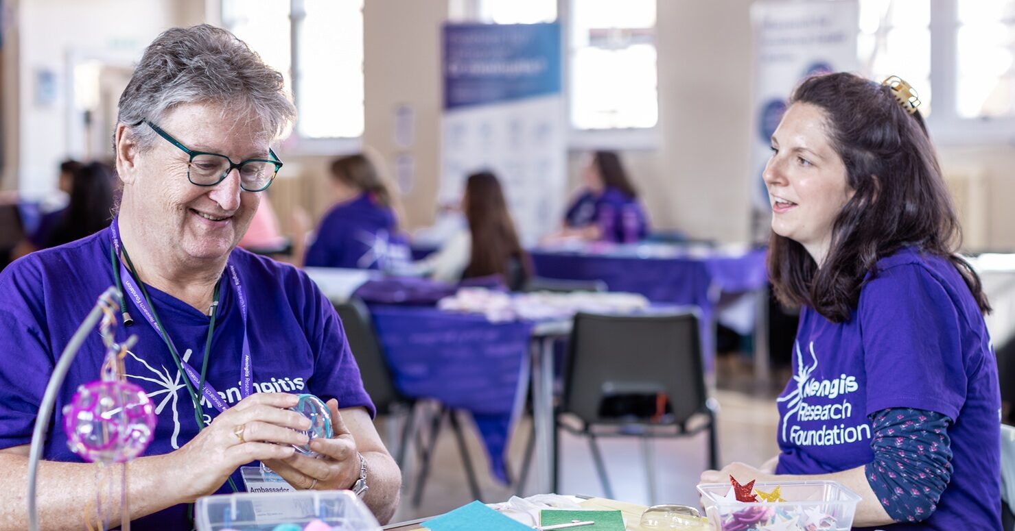 A man and a woman sat at a table together whilst the man is crafting. The woman is talking and the man is smiling.