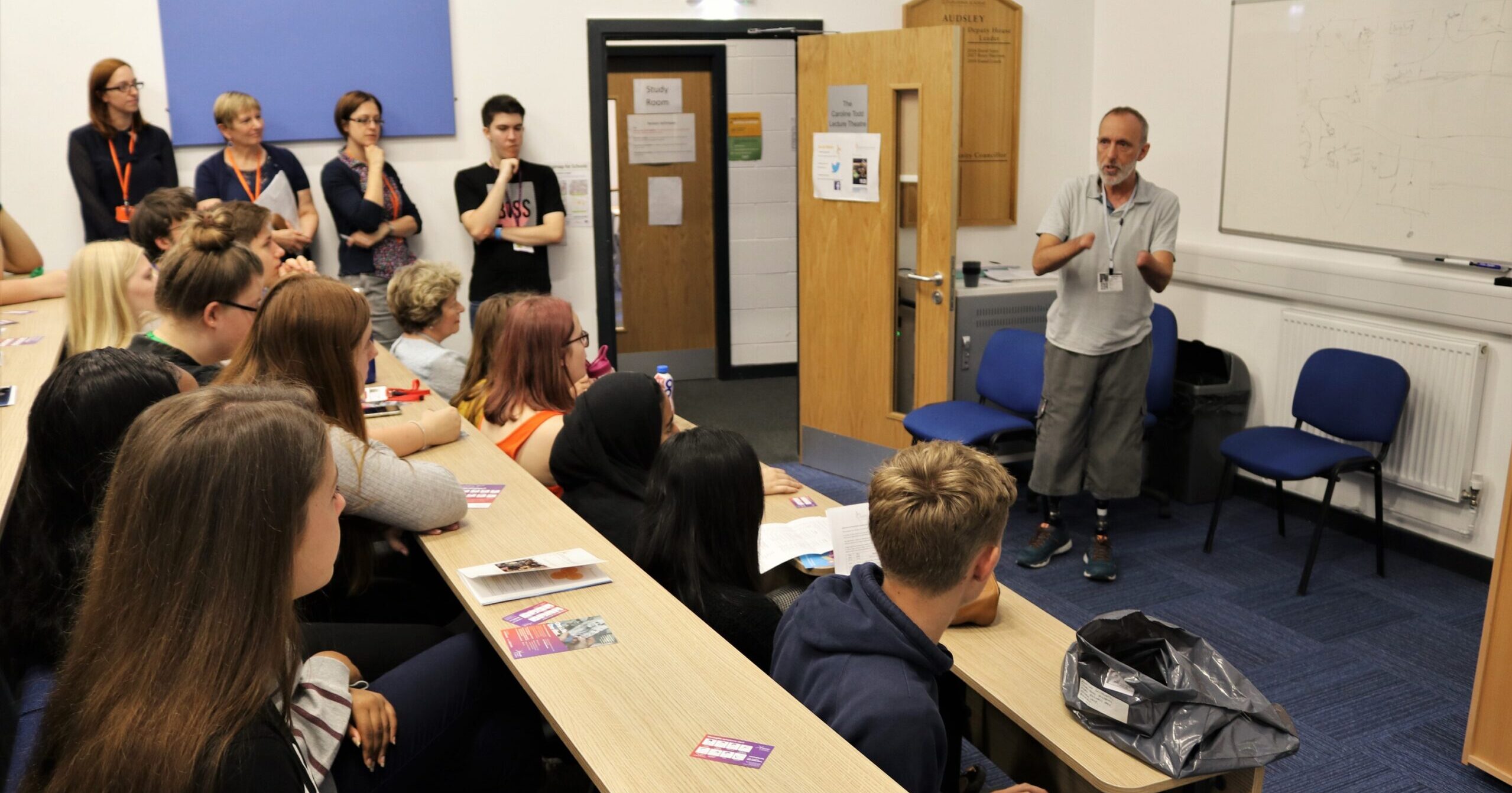 Classroom of teenagers sat at tiered desks listening to a man who has survived meningitis talking about his experience. Man is at the front of the teenagers standing up and showing his prosthetic legs.
