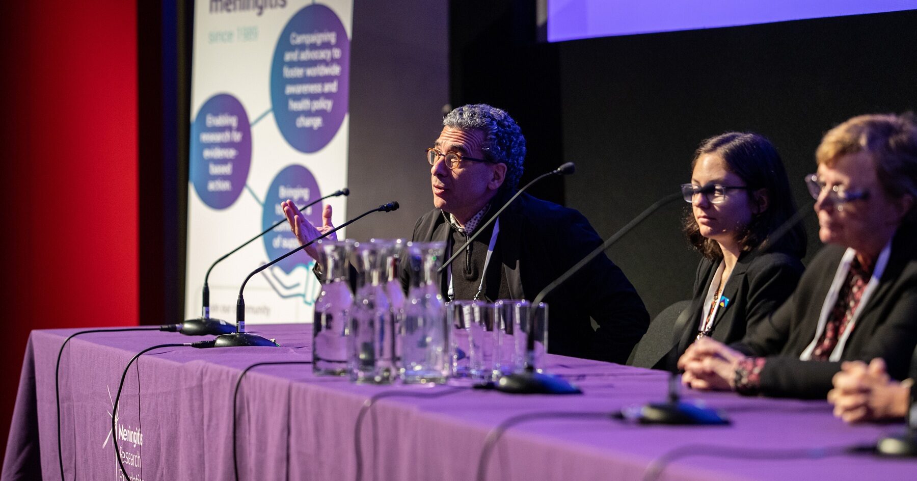 Man at an event, sat at a table speaking into a microphone, with two women sat next to him.
