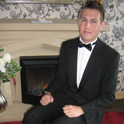 A photograph of Jake Hilton in a tuxedo seated by a fireplace