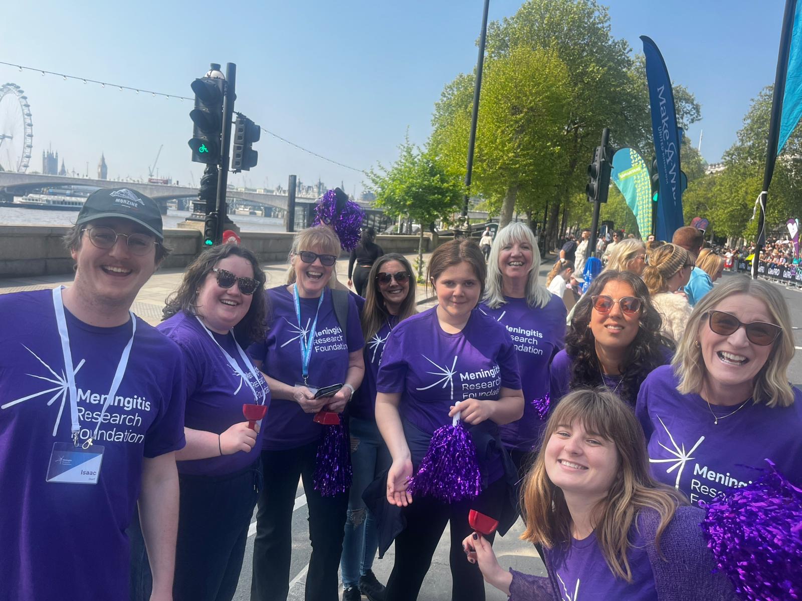 Nine people wearing Meningitis Research Foundation t-shirts smiling and holding pom poms.