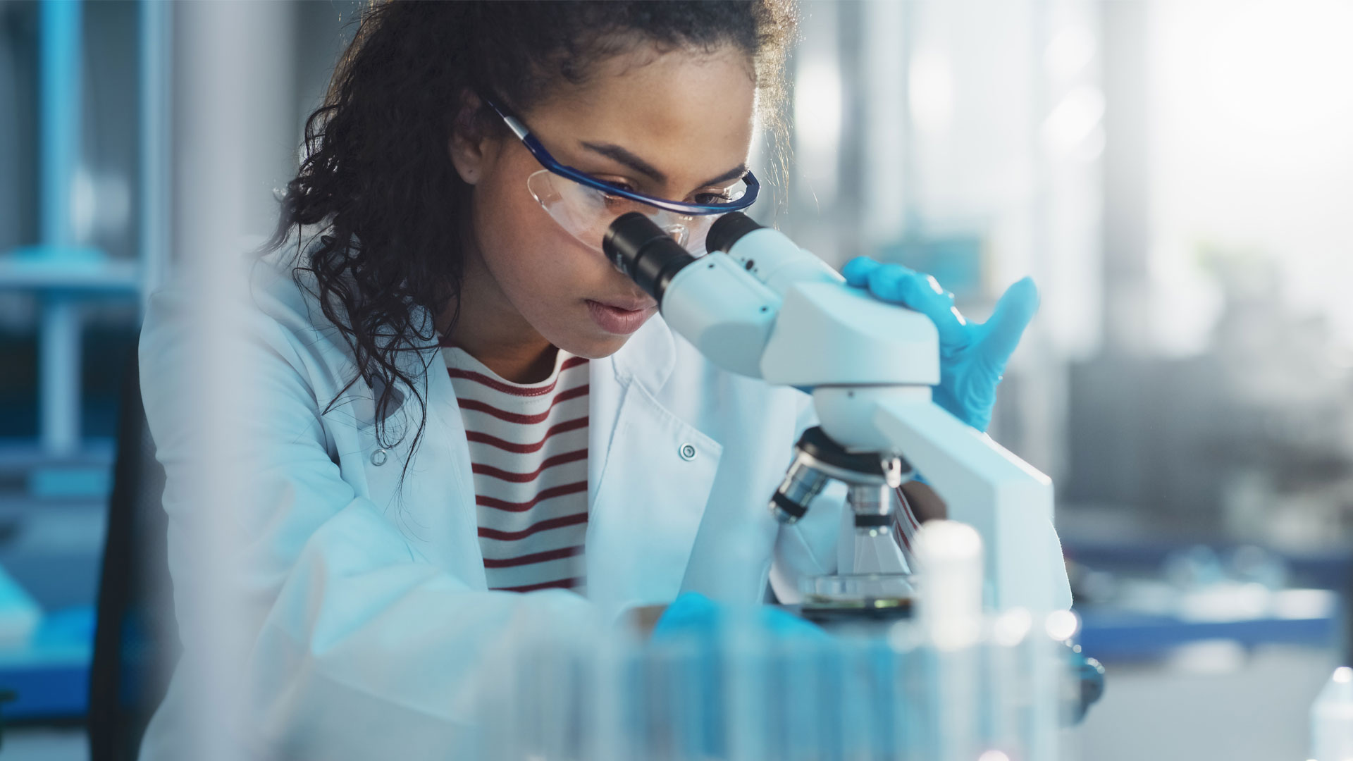 A scientist wearing goggles and a lab coat looking into a microscope.