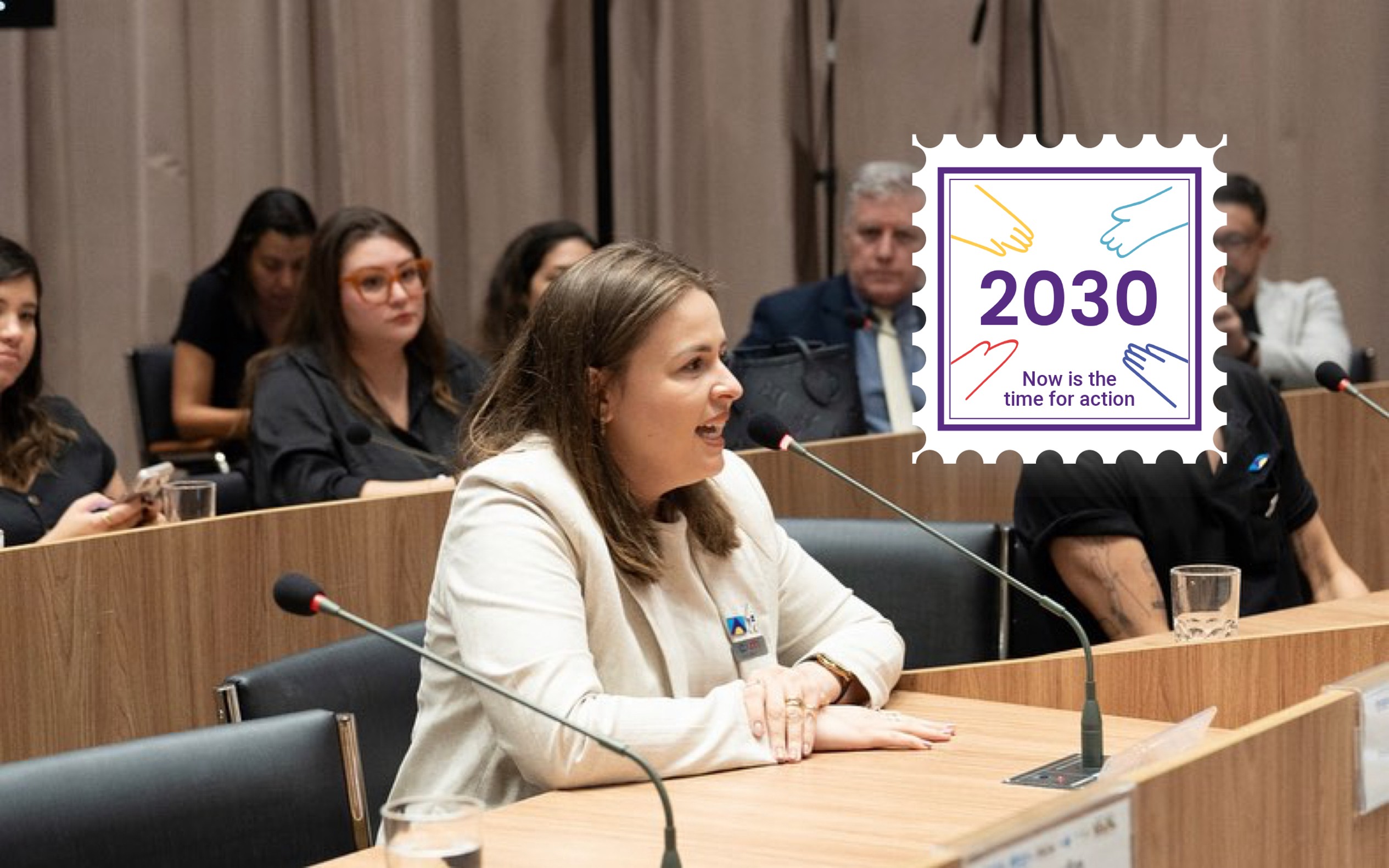 Suelen, a Brazilian meningitis advocate, speaking at an official meeting. There is a campaign badge in the corner of the image which looks like a stamp and says 