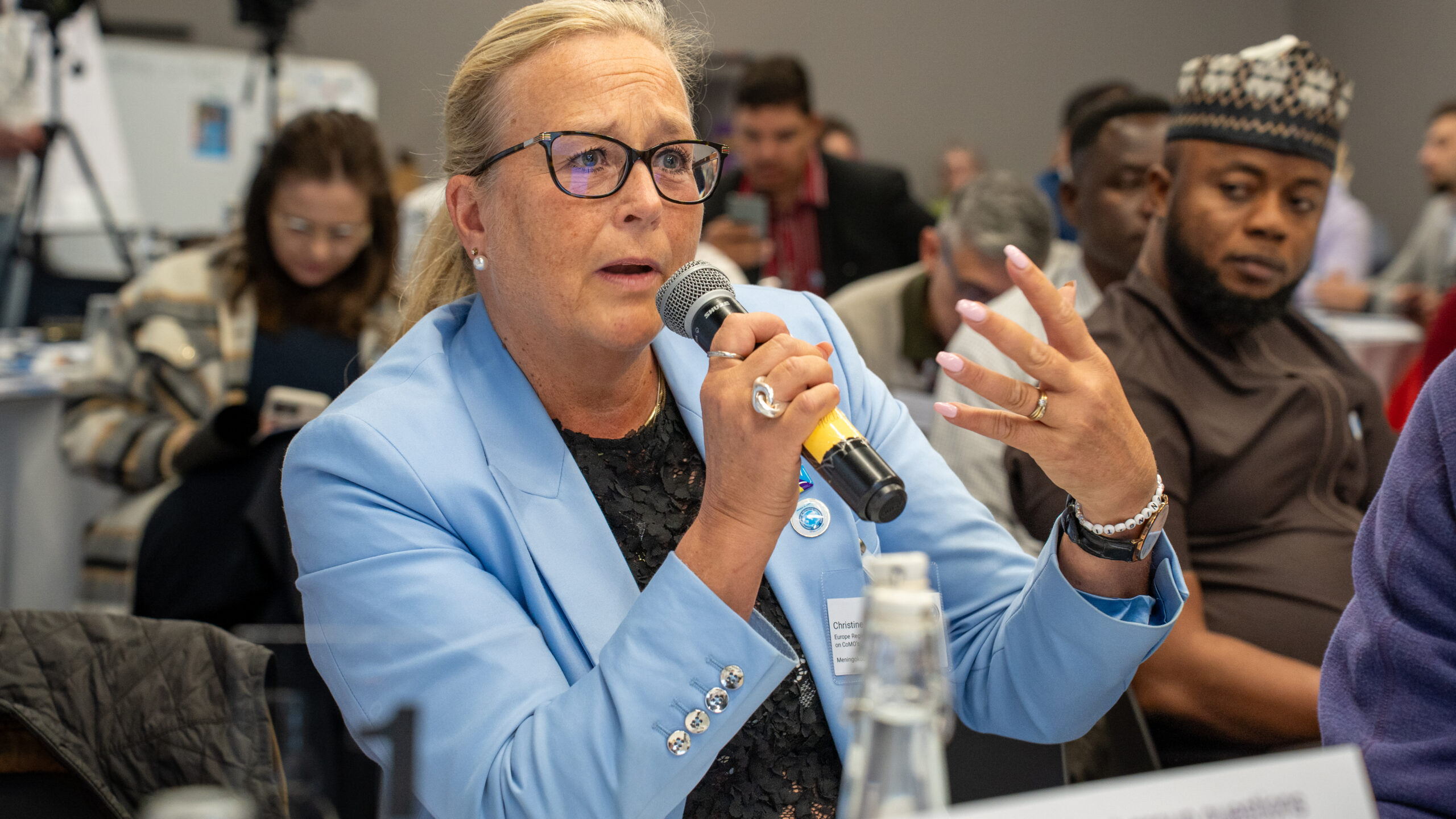 A woman speaking into a microphone being watched by various CoMO members.