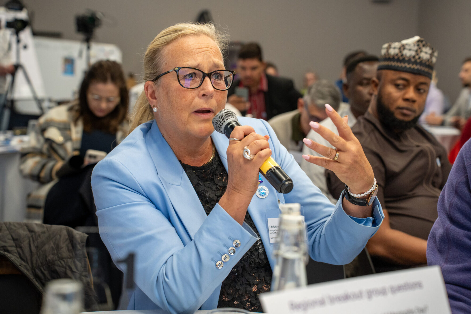 A woman speaking into a microphone being watched by various CoMO members.