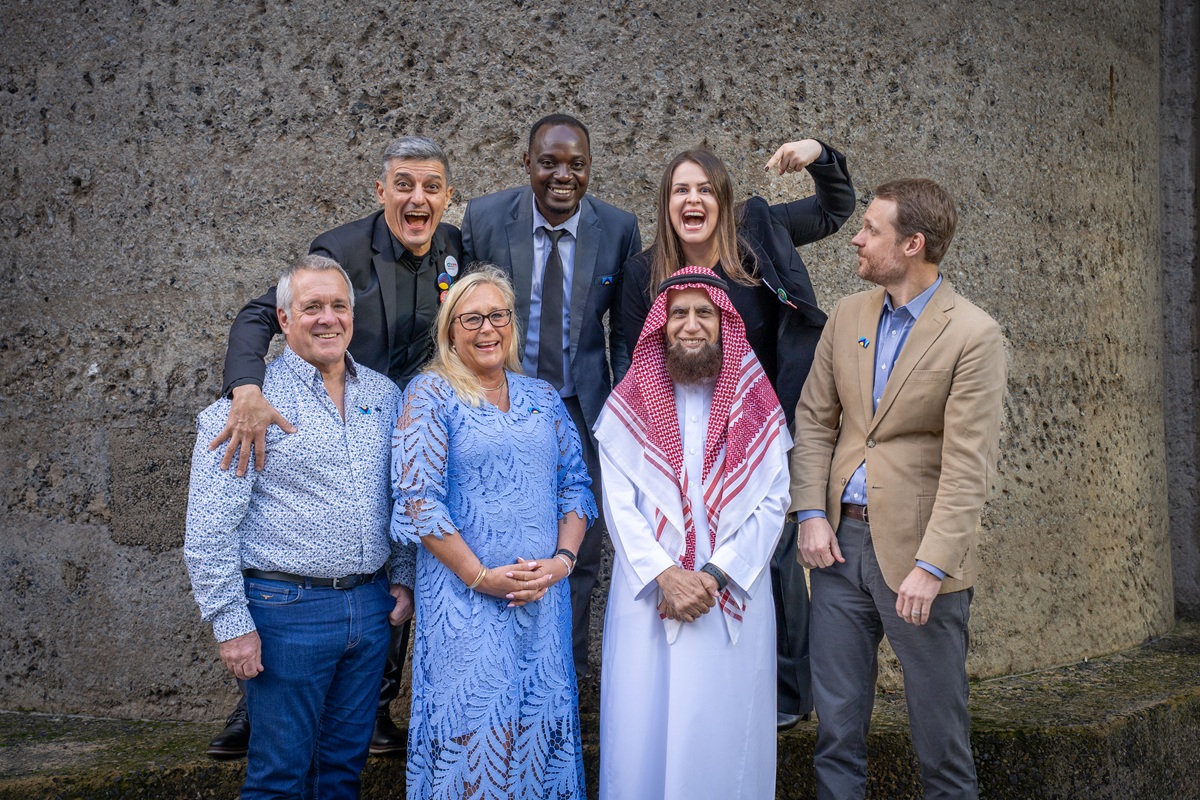 Group shot of seven people standing together, laughing and smiling against a stone wall.