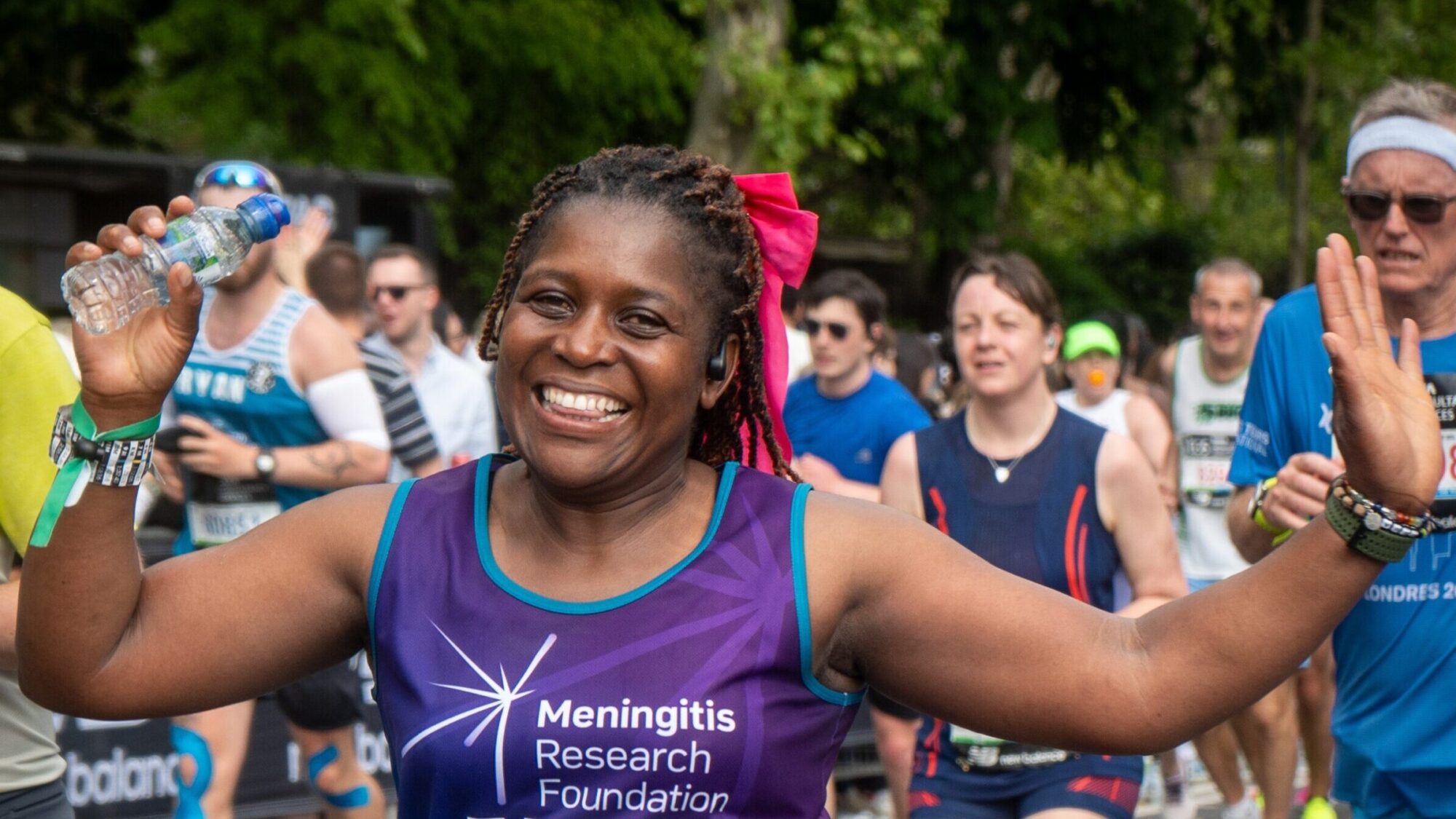A London Marathon runner with a bow in her hair waves her hands in the air and smiles for the camera.