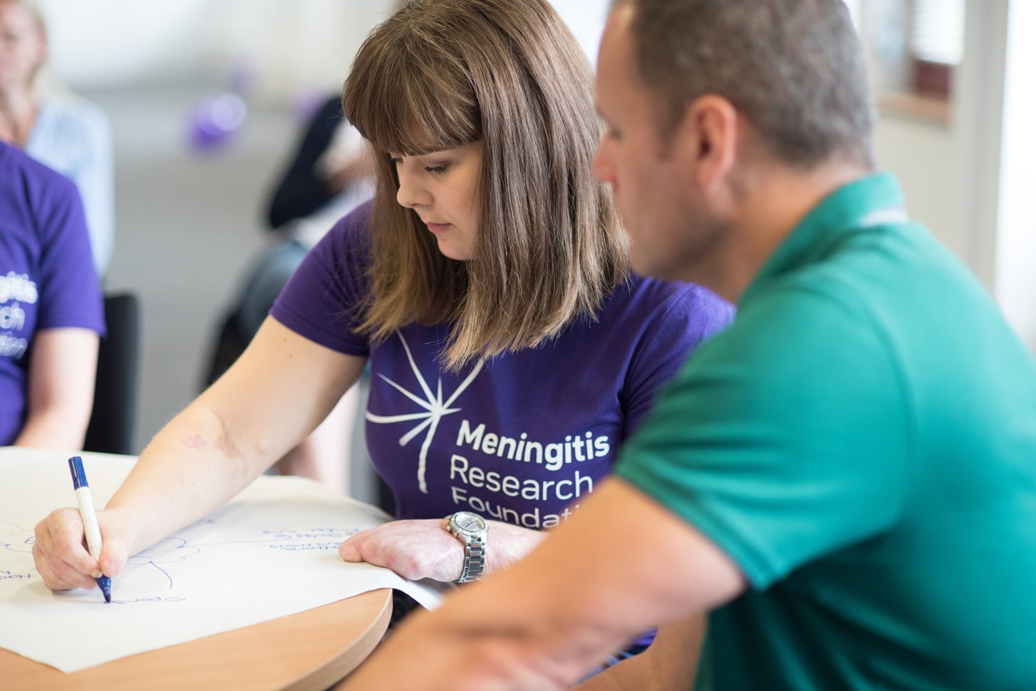 A woman in a Meningitis Research Foundation t-shirt sitting at a table with a man in a meeting room. She is writing on a flipchart.