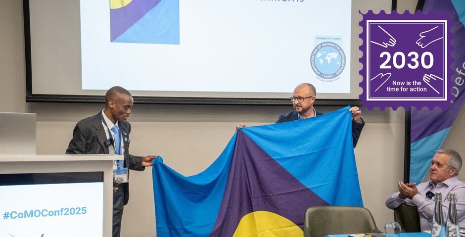 Two men holding up the Meningitis Flag at the CoMO members' conference 2025.