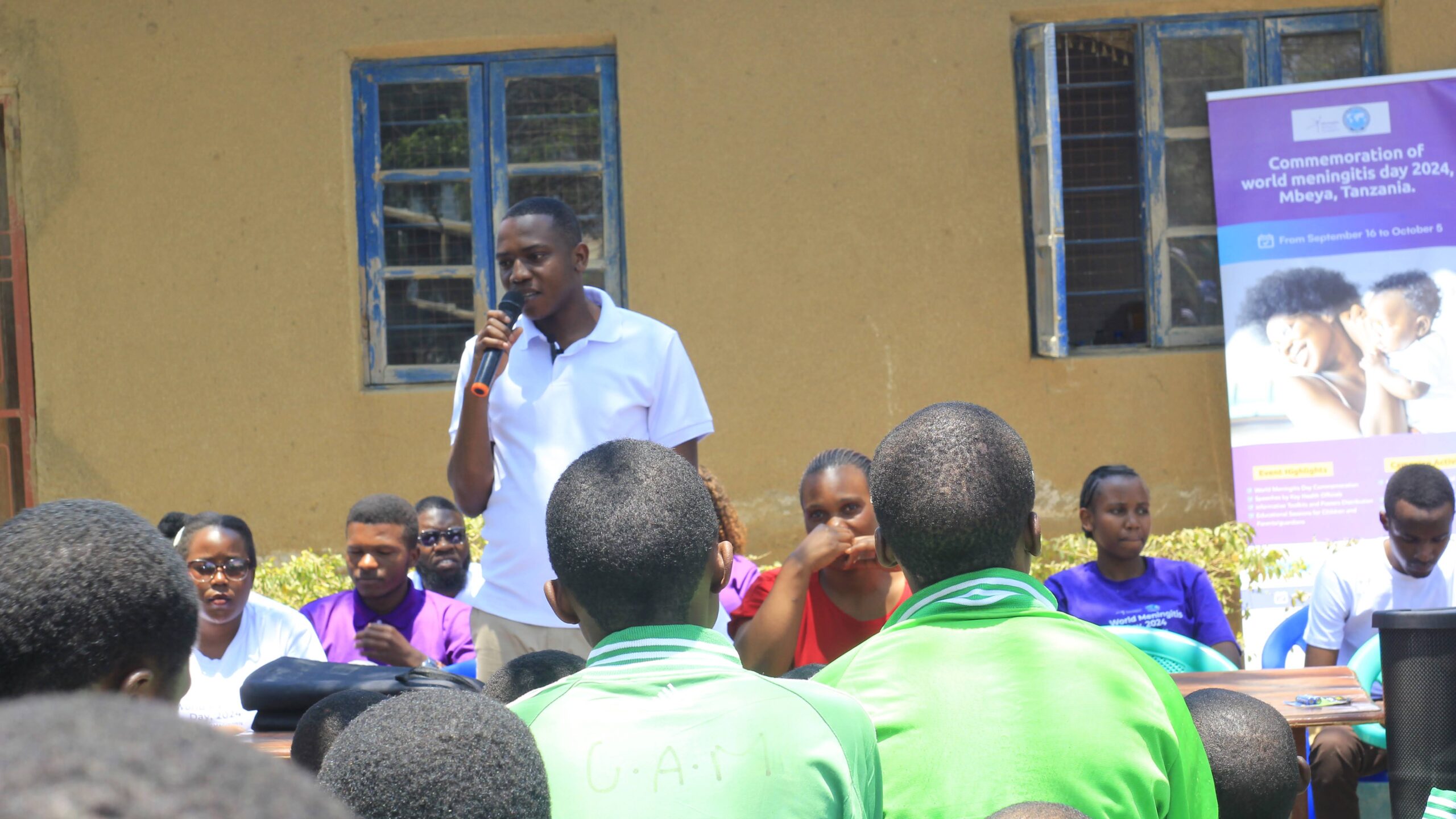 A man speaking into a microphone to a crowd sitting outside.