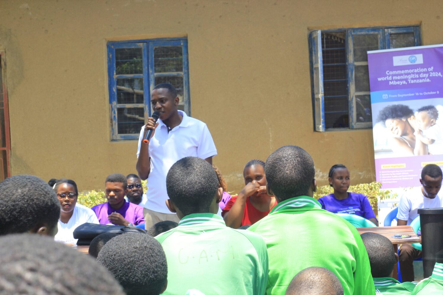 A man speaking into a microphone to a crowd sitting outside.