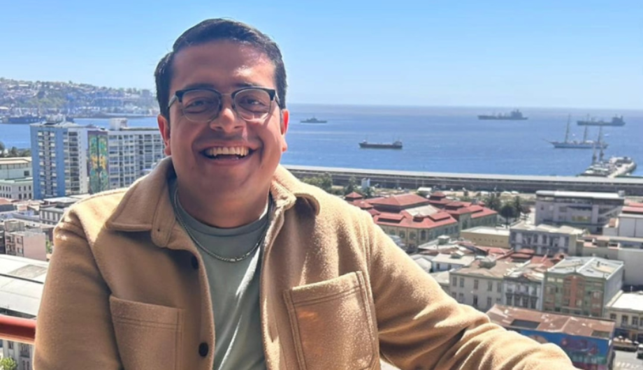 A young man wearing glasses smiling in front of a city backdrop.