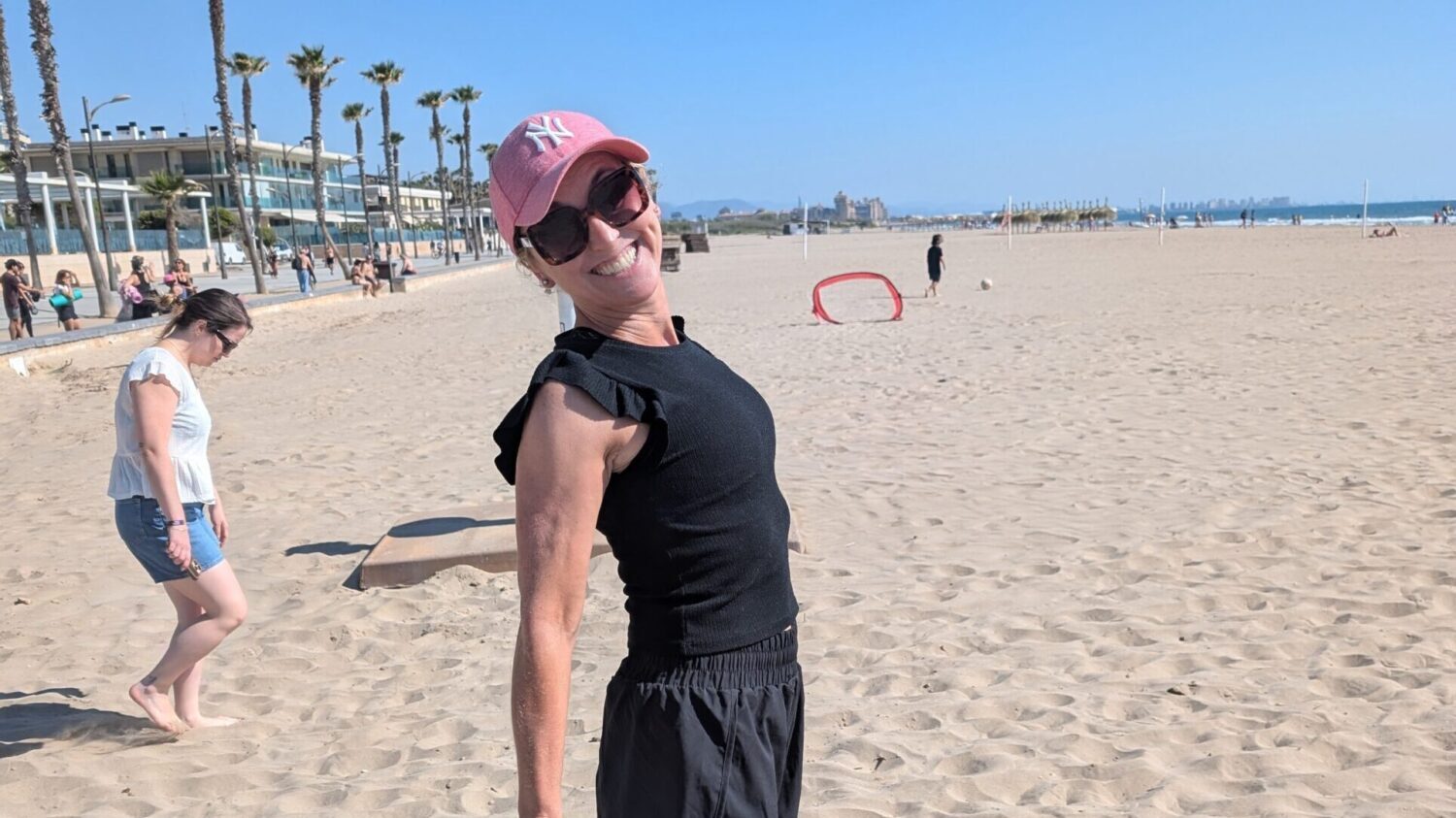 A woman with sunglasses and a pink baseball cap smiling on the beach.