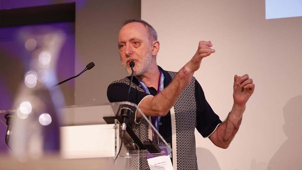 A man speaking at a lectern. He wears a Meningitis Research Foundation lanyard and is gesticulating with his forearms, which have surgical scars.