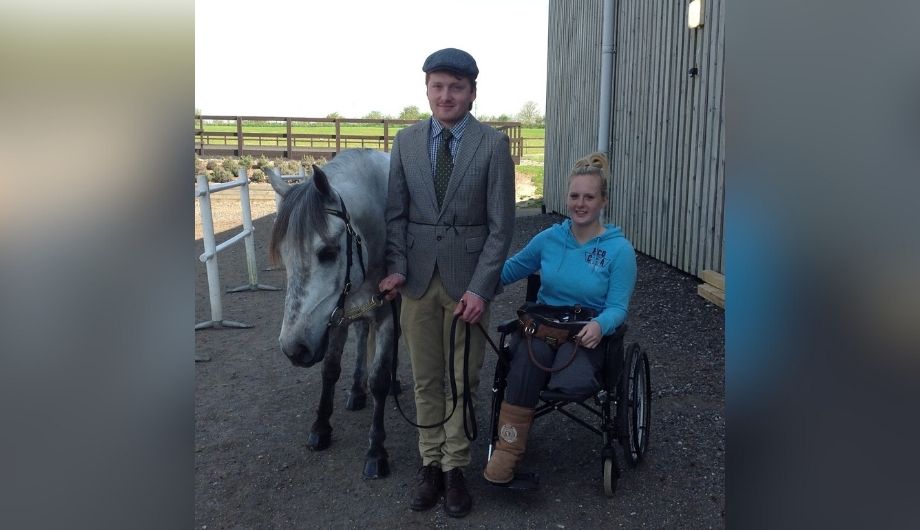 A blonde woman wearing a bright blue hoodie, in a wheelchair holding hands with a smartly-dressed man standing next to a horse.