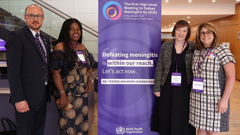 Four people smiling and standing next to a banner that reads: 'Defeating meningitis is within our reach. Let's act now.'