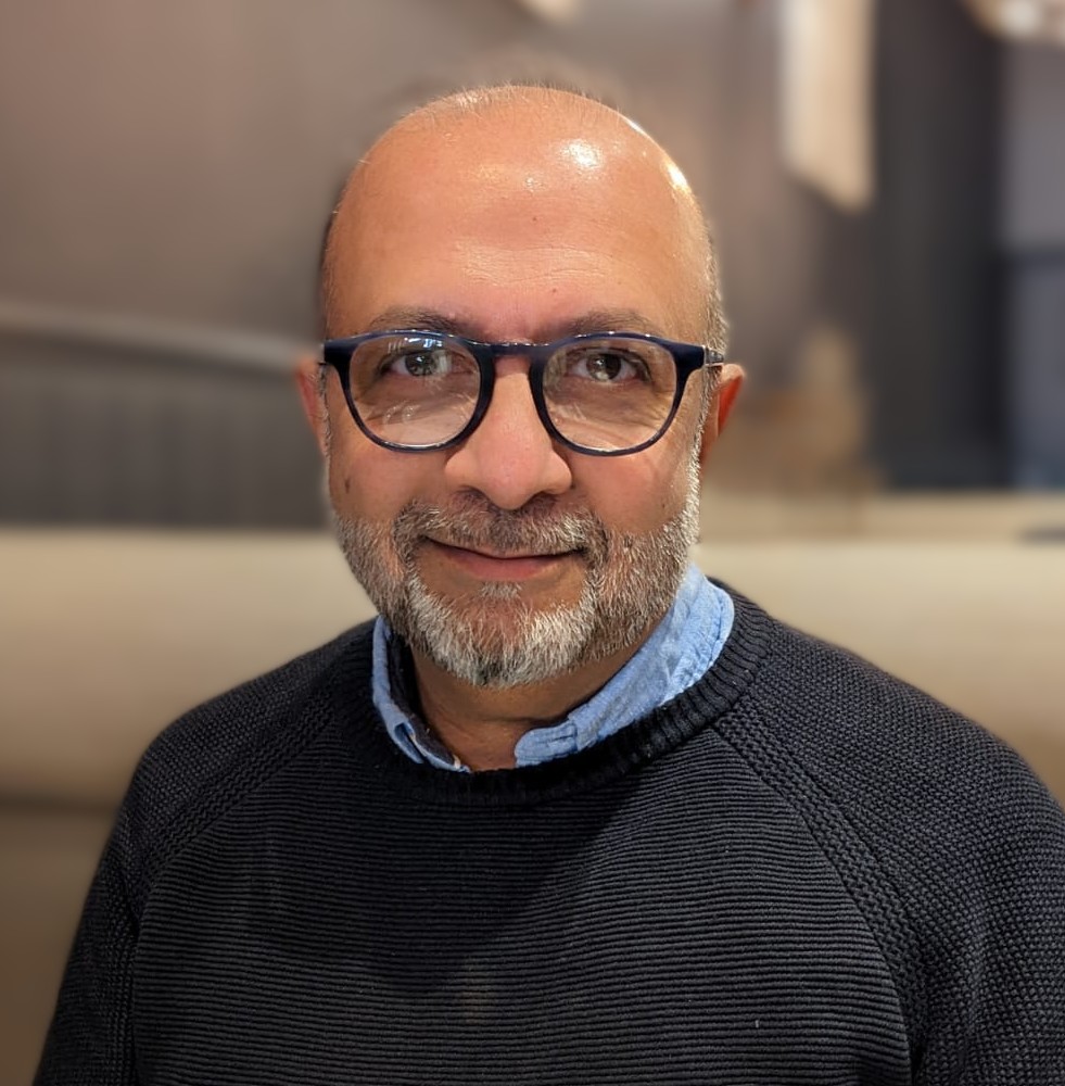 Man with glasses and a beard, wearing a blue shirt and black jumper, looking into the camera with a blurred background behind him.
